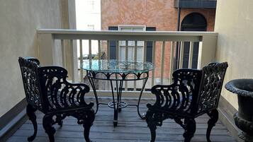 A balcony with two ornate metal chairs and a glass table, overlooking a building with a blue awning and a door.