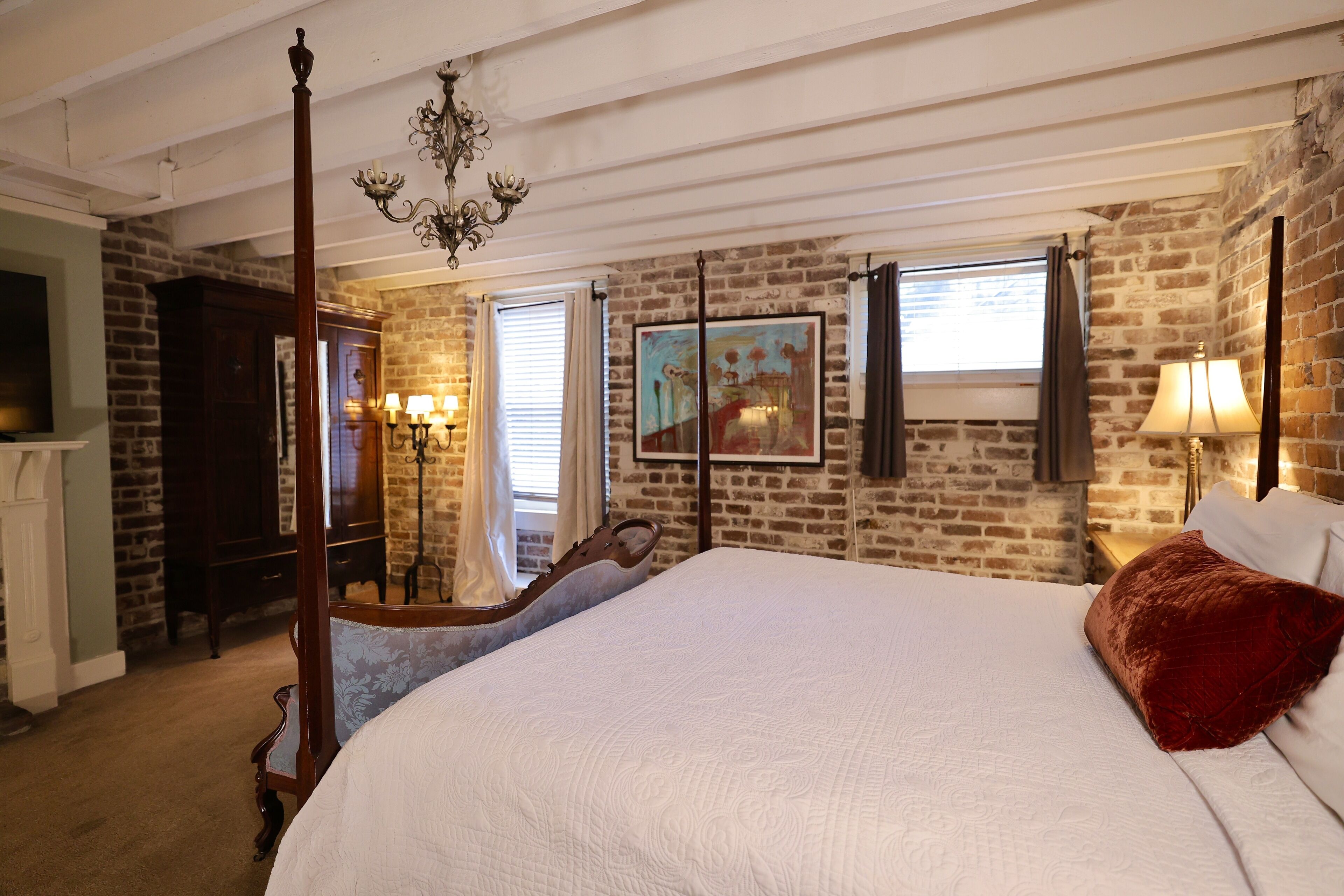 A bedroom with a four-poster bed, a chandelier, a large painting, and a brick wall.