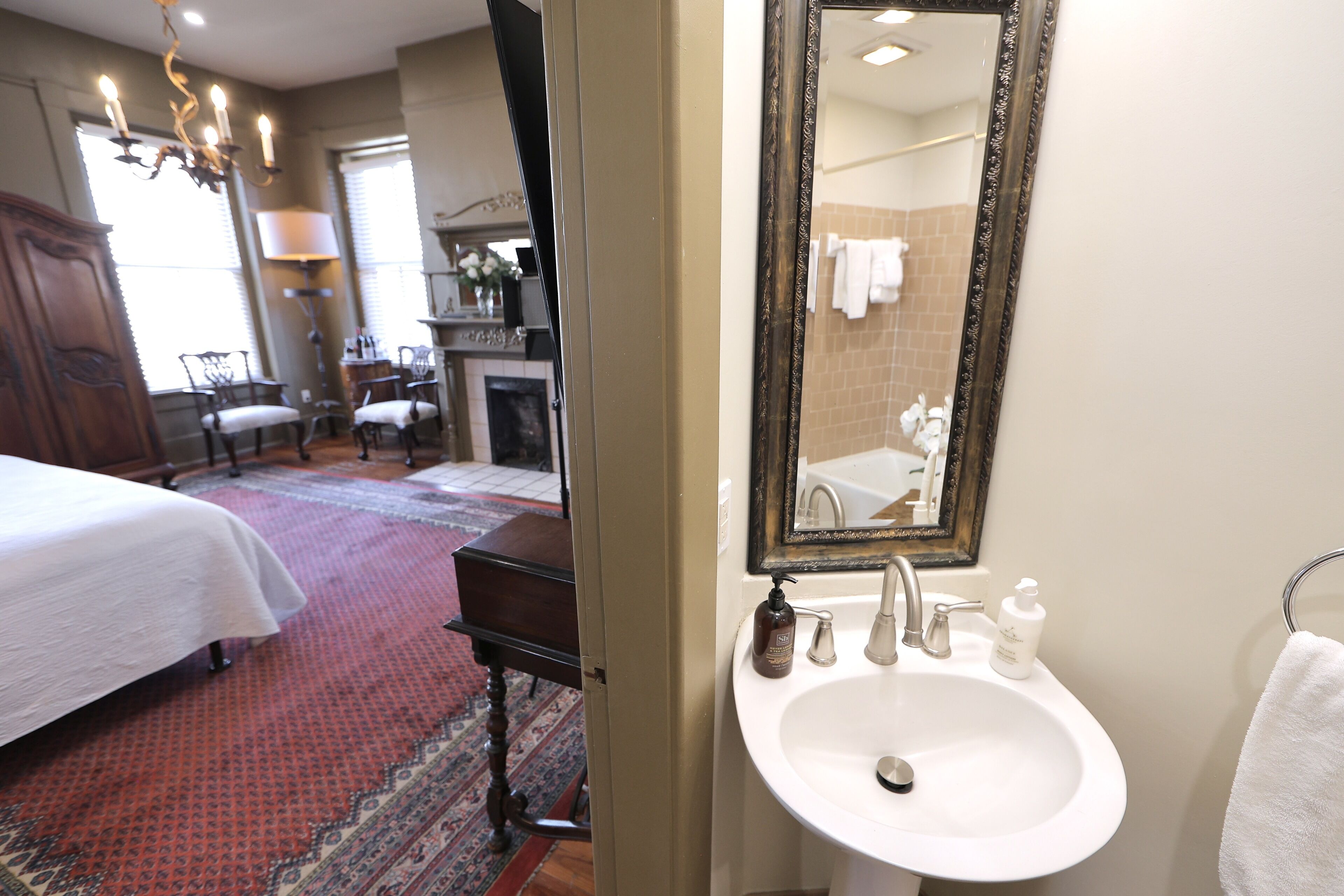 Una habitación de hotel con cama, zona de estar con sillas, espejo grande, lavabo blanco y baño con bañera y toallero.