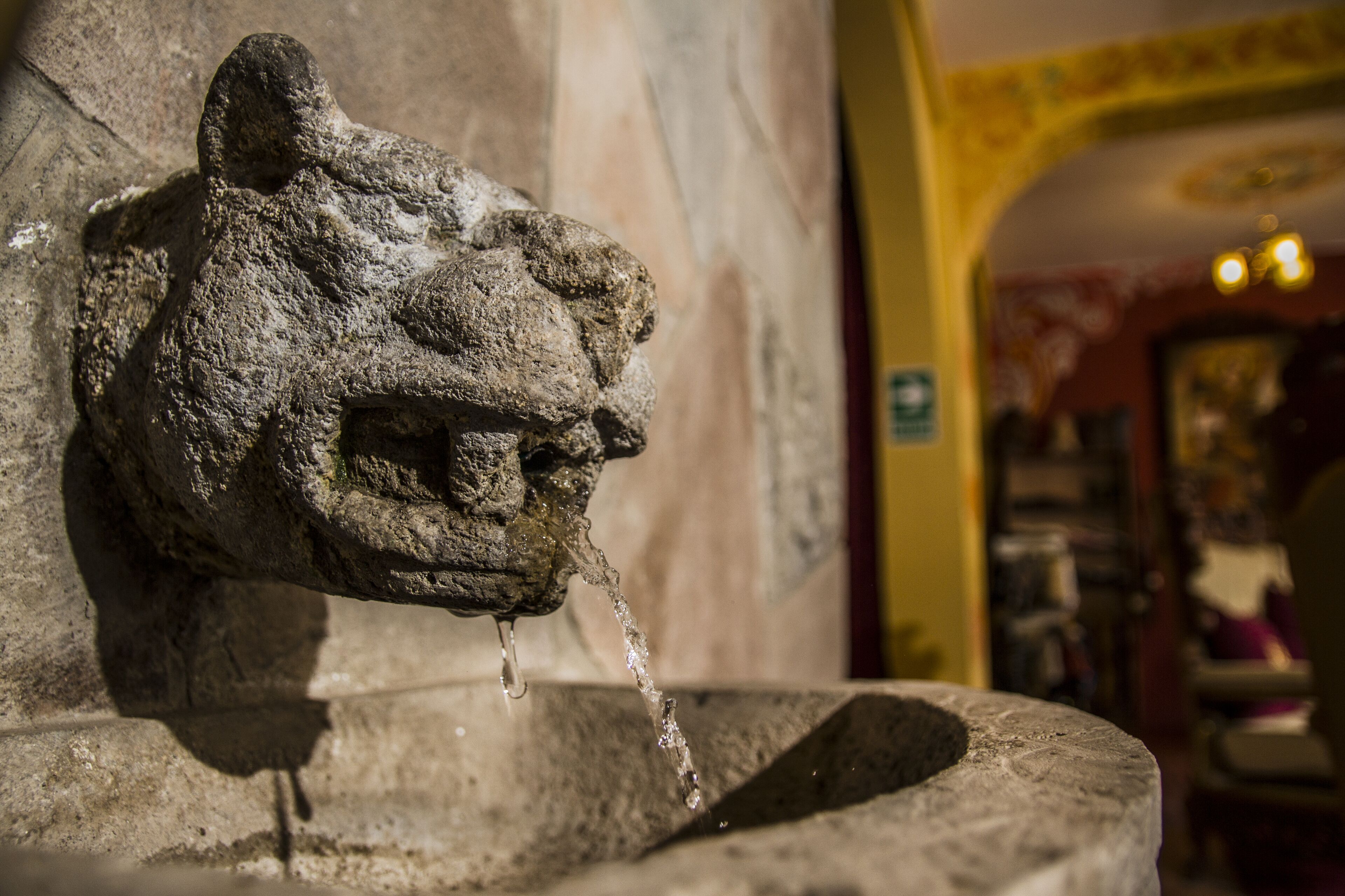 A stone water spout with water flowing from it, set against a wall with a yellow archway in the background.