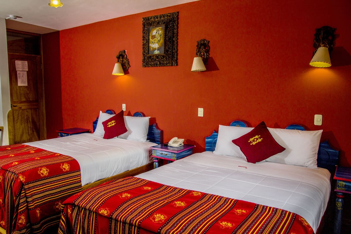 Habitación de hotel con dos camas, ropa de cama roja y blanca, un cuadro en la pared y dos lámparas de pared.