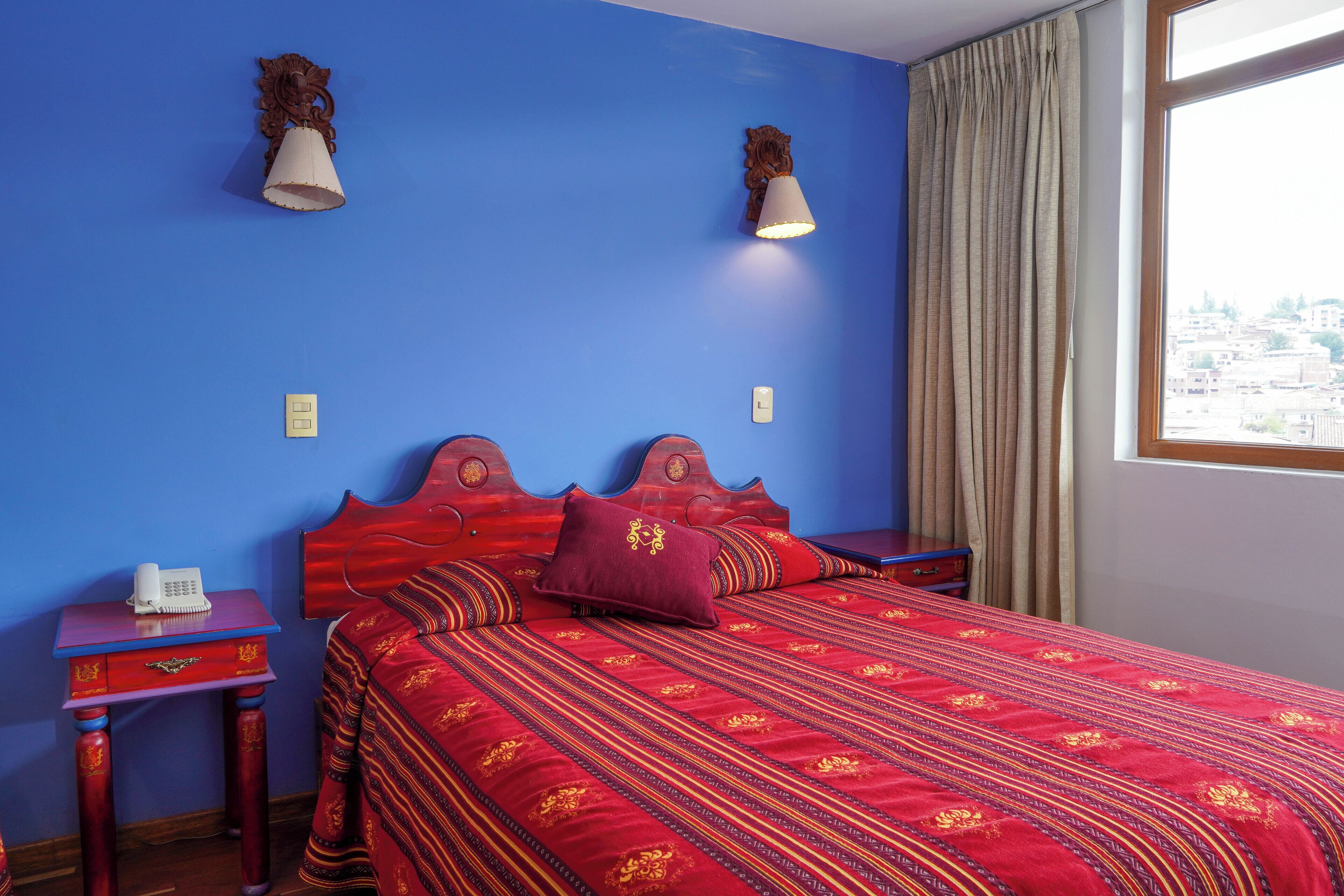A hotel room with a red bedspread, two bedside lamps, a wooden nightstand with a phone, and a window with a view of buildings.