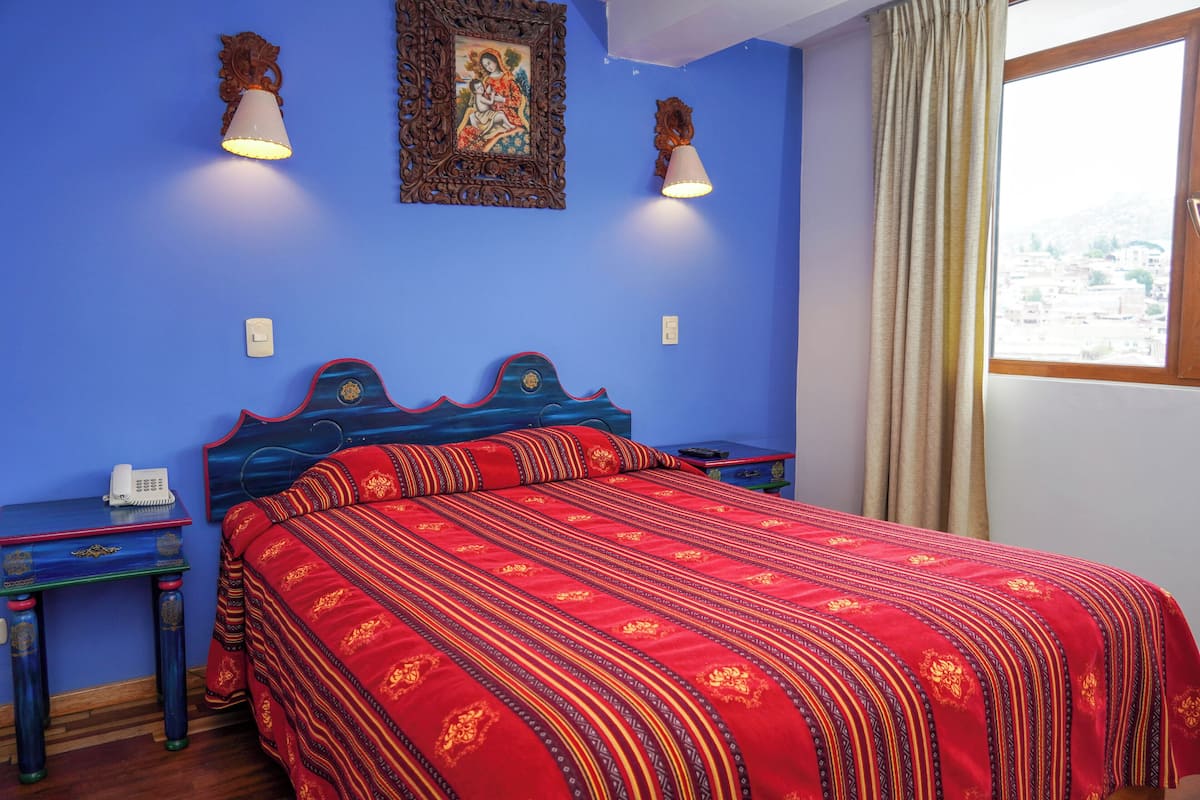 Una habitación con una pared azul, una cama con colcha estampada roja, dos mesitas de noche con lámparas y un cuadro enmarcado sobre la cama.