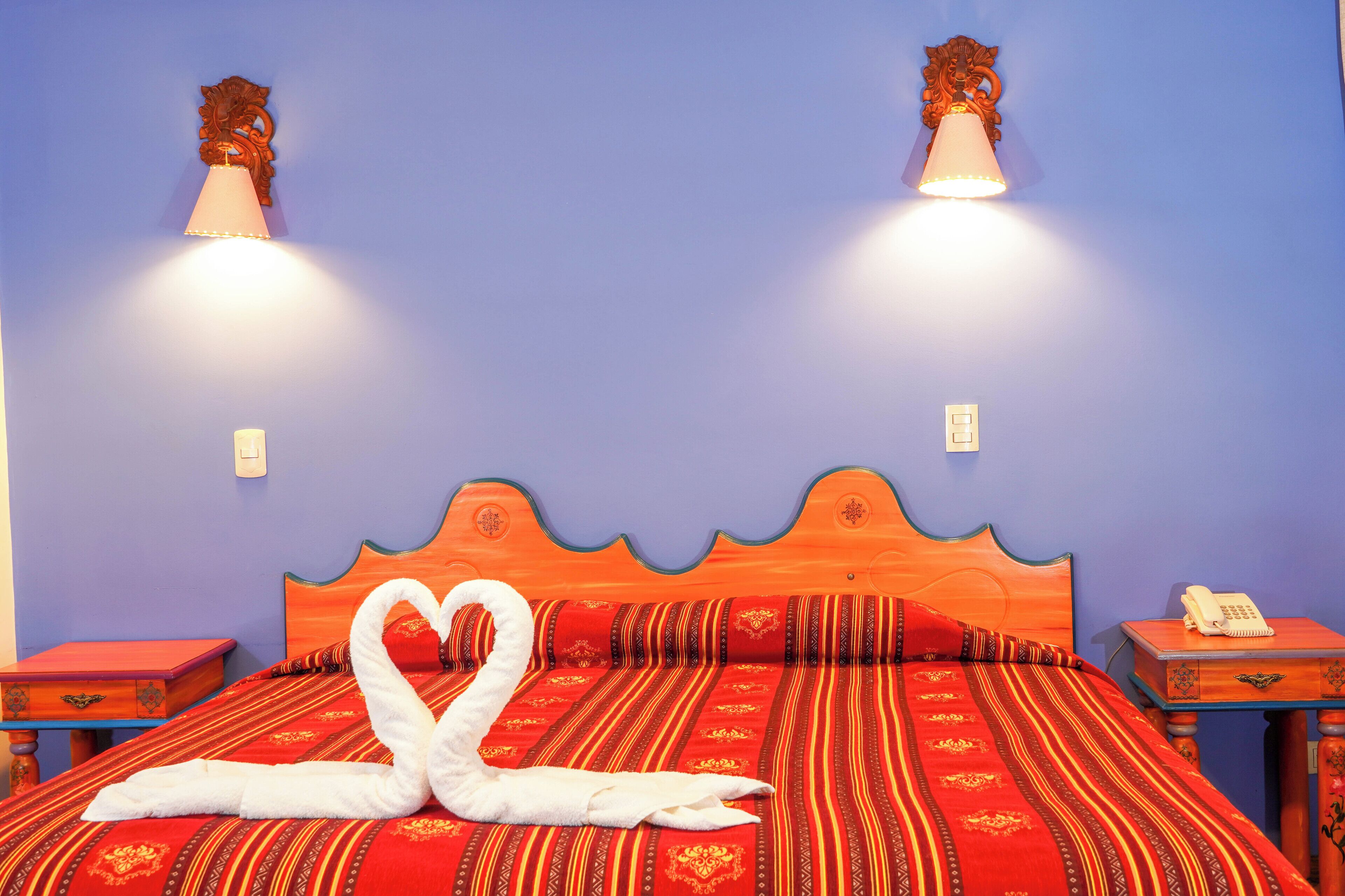 A bed with a red and orange patterned bedspread, two wall-mounted lamps, and a swan-shaped towel arrangement.