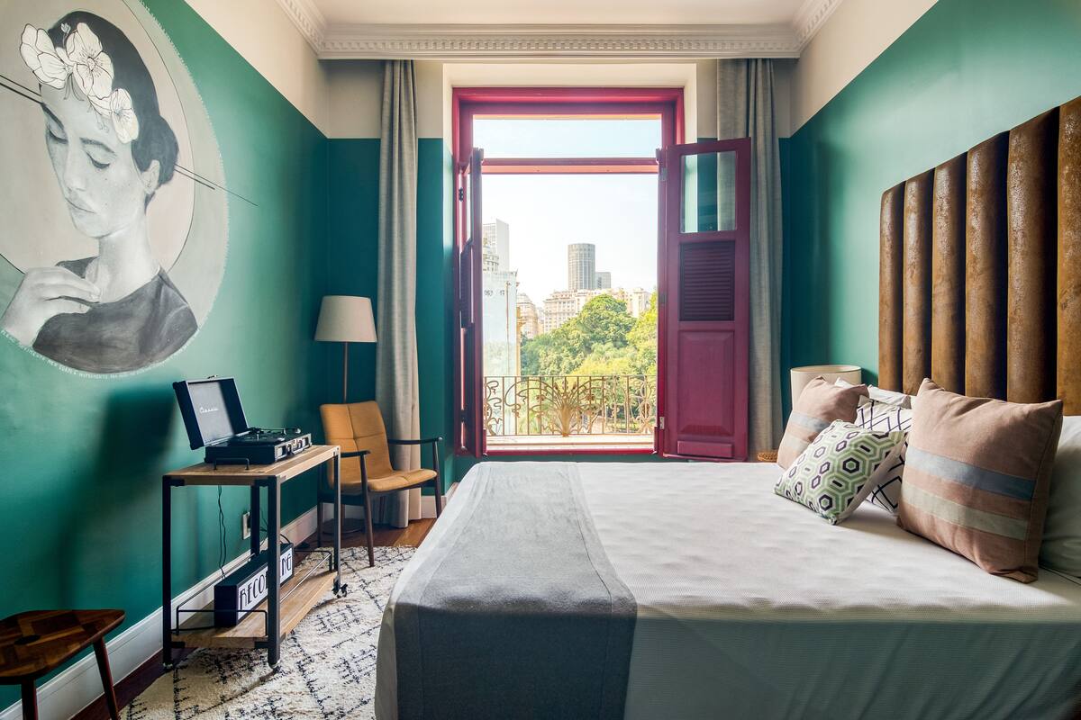 A hotel room with a bed, a desk with a laptop, a chair, a window with a view of buildings and trees, and a wall with a mural.