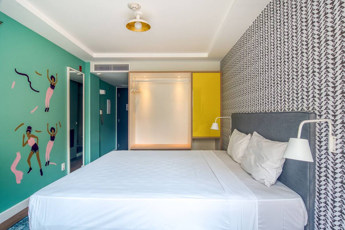 A hotel room with a large bed, a wardrobe, a bathroom, and a wall with a geometric pattern.
