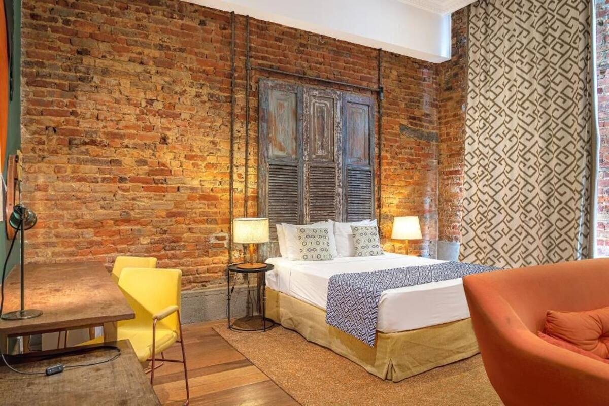 A room with a brick wall, a bed with a patterned blanket, a yellow chair, and a small table with a lamp.