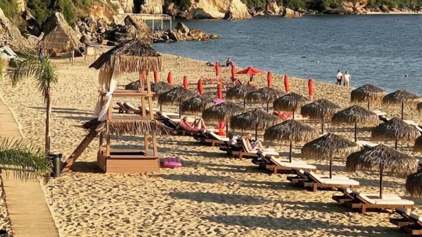 Una playa con tumbonas y sombrillas, una estructura de madera y vistas al mar y colinas.