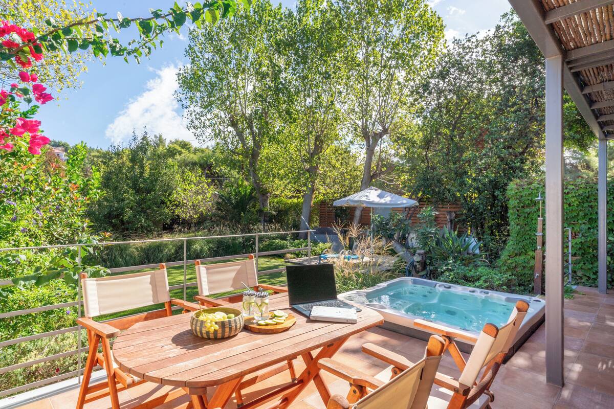 A wooden table with chairs, a laptop, and a bowl of fruit on a patio with a hot tub and lush greenery in the background.