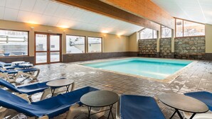 An indoor swimming pool with lounge chairs and tables, stone walls, and large windows.