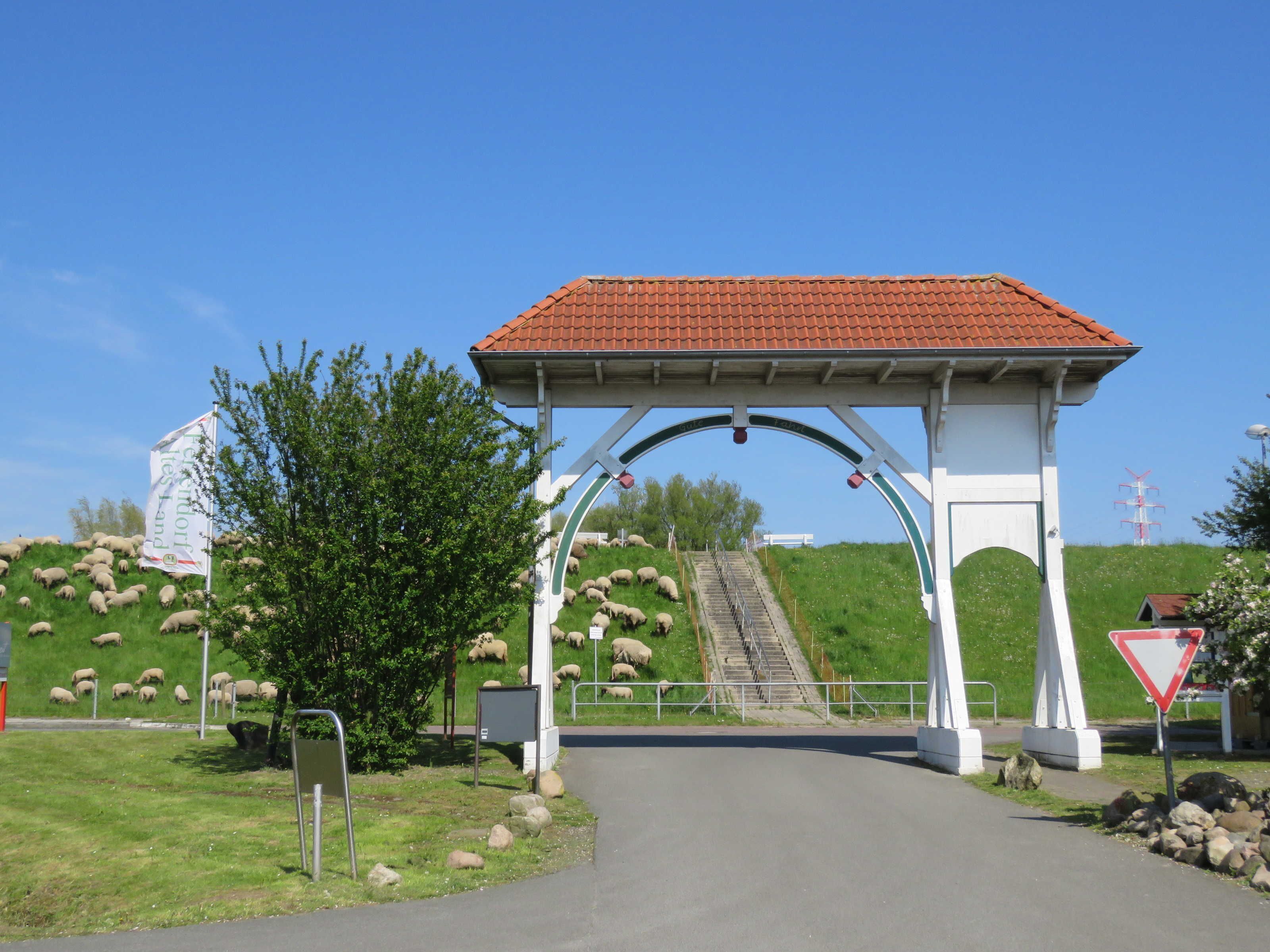 En lantlig entré med en vit valvformad öppning, ett rött tegeltak och en grönskande kulle med får.