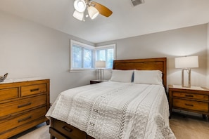 Une chambre avec un lit en bois, une table de chevet avec une lampe, une commode avec des tiroirs, un ventilateur de plafond et une fenêtre avec des stores.