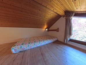 A room with a sloped ceiling, wooden flooring, a bed with a patterned bedspread, a window with curtains, and a wall-mounted light fixture.