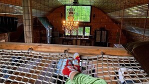 A hammock suspended in a rustic wooden room with a large window, a chandelier, and a wooden table.