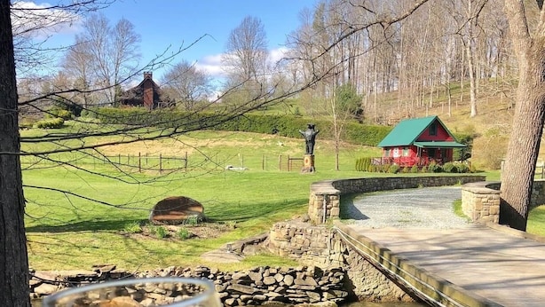 A glass of red wine held in front of a rural landscape with a stone bridge over a stream, a red building, and a statue in the background.
