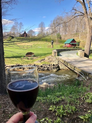 A glass of red wine held in front of a rural landscape with a stone bridge over a stream, a red building, and a statue in the background.