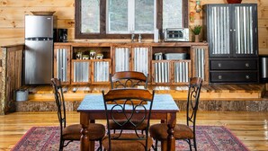 A rustic wooden interior with a dining table, chairs, a chandelier, and a kitchen area.