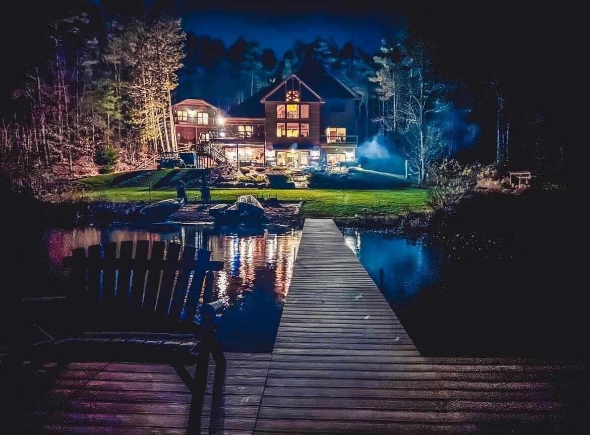 A large house with a dock extending into a calm body of water, surrounded by trees.