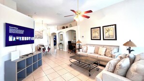 A spacious living room with a large flat-screen TV mounted on the wall, a ceiling fan, a coffee table, a sofa, and a TV stand with shelves.