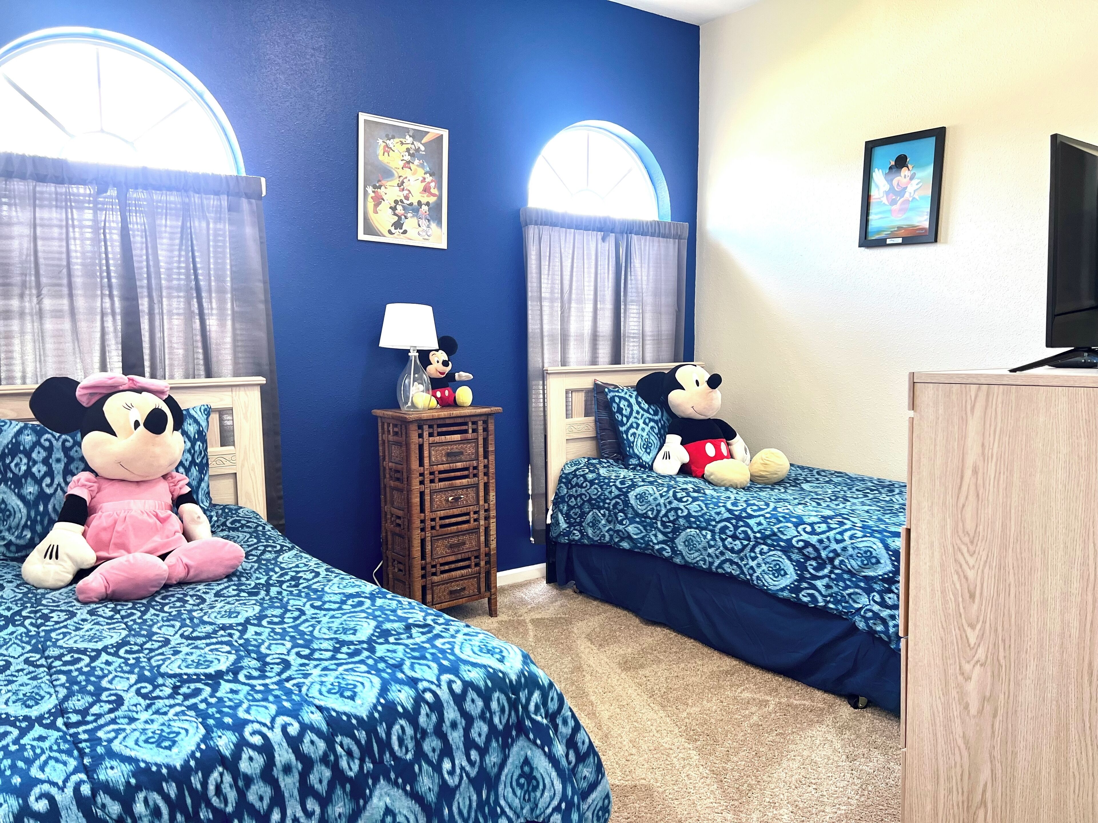 Quarto com duas camas, cada uma com um brinquedo de pelúcia do Mickey Mouse. Há uma mesinha com um abajur e um porta-retrato na parede.