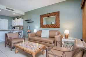 Un salon avec un canapé en cuir marron, deux fauteuils, une table basse surmontée d’un plateau en mosaïque, une cuisine à l’arrière-plan et un grand miroir accroché au mur.