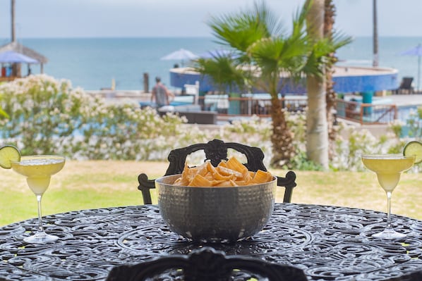 Une table avec un bol de chips et deux boissons, le tout placé devant une vue sur une piscine et des palmiers.