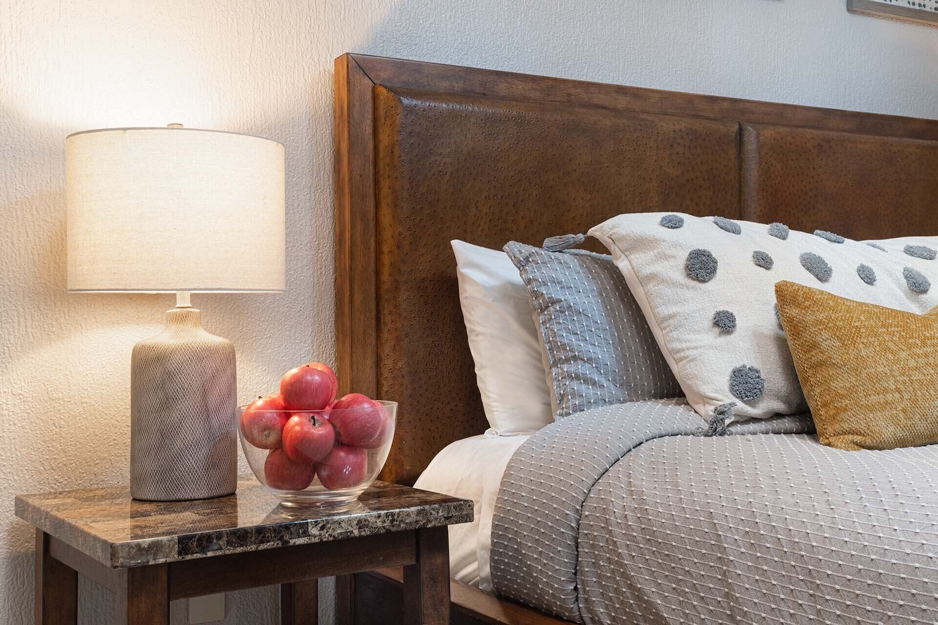 Une chambre à coucher bien rangée, avec une tête de lit en bois, une table de chevet sur laquelle se trouve une lampe, et un bol de pommes.