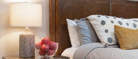 Une chambre à coucher bien rangée, avec une tête de lit en bois, une table de chevet sur laquelle se trouve une lampe, et un bol de pommes.