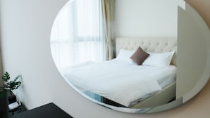 Un espejo circular refleja una cama bien hecha con ropa blanca y un cabecero capitonado, una ventana con cortinas de encaje y una pequeña planta en una mesita de noche.