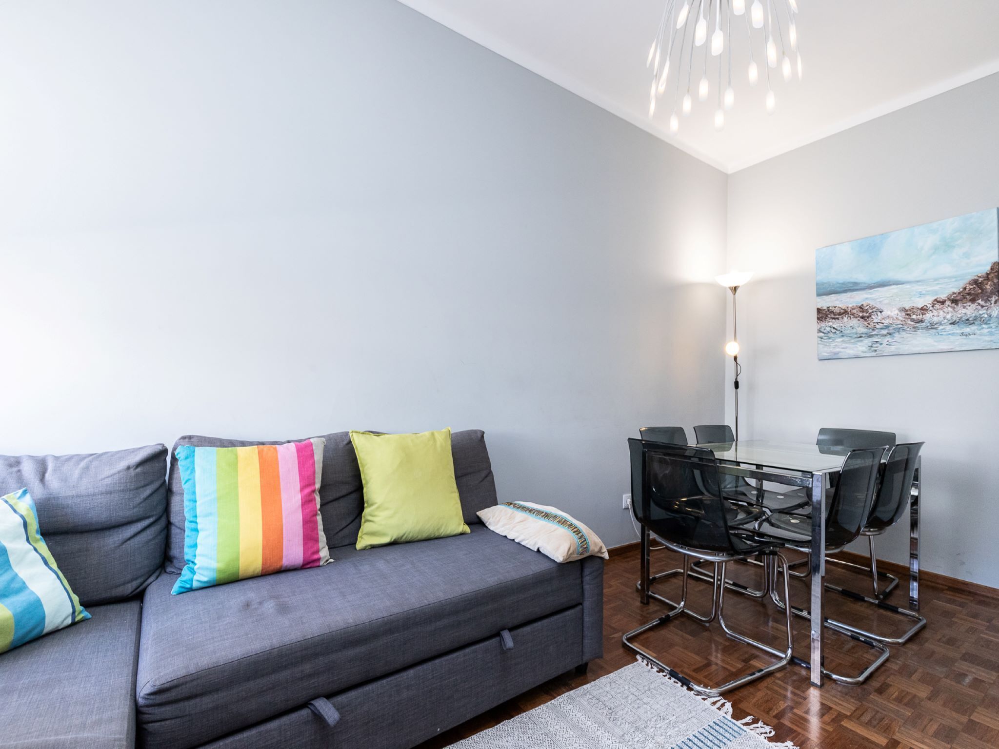 Ein modernes Wohnzimmer mit einem grauen Sofa, farbenfrohen Kissen, einem Glastisch mit Stühlen, einer Stehlampe und einem Gemälde an der Wand.