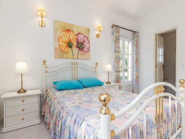 A bedroom with a bed, two bedside lamps, a nightstand, a dresser, a floral painting, and a window with curtains.