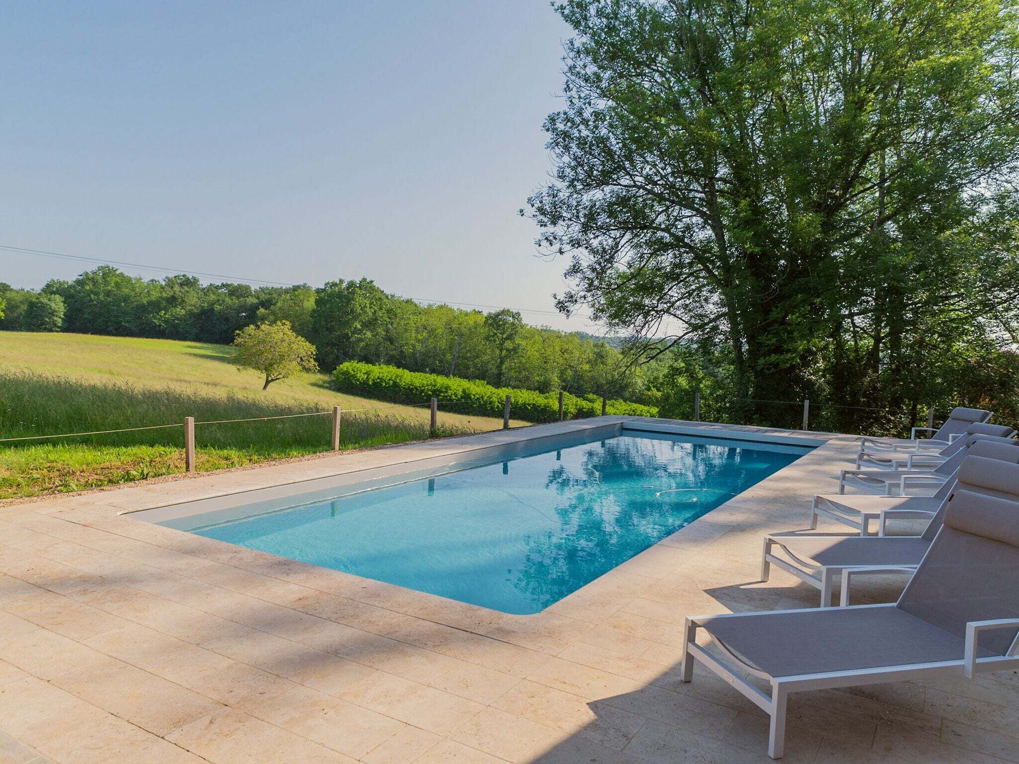 A rectangular swimming pool with a clear blue water surface, surrounded by a paved deck. There are several lounge chairs with cushions arranged around the pool.