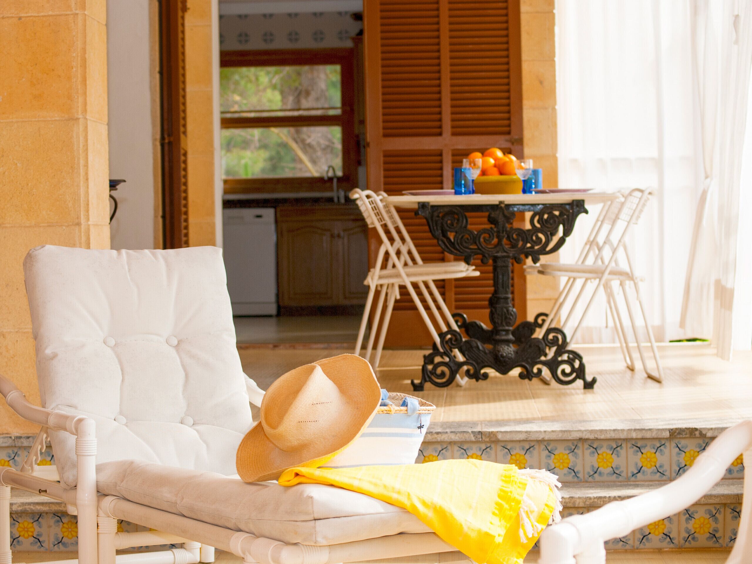 A sunlit room with a white cushioned chair, a table with oranges, and a view of a kitchen and greenery outside.