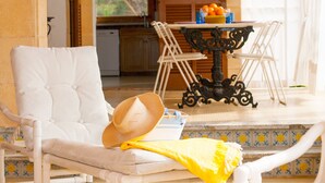 A sunlit room with a white cushioned chair, a table with oranges, and a view of a kitchen and greenery outside.