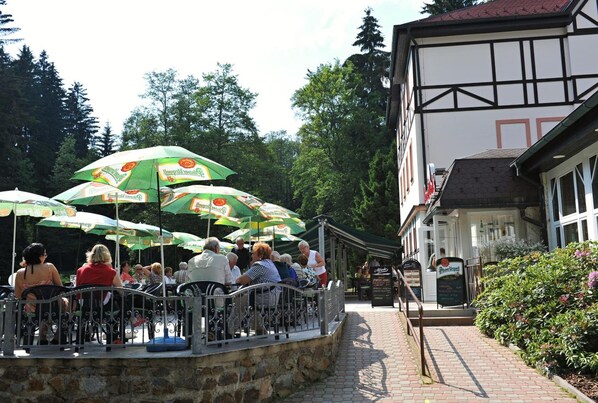An outdoor seating area with tables and chairs, surrounded by greenery and a building with a traditional architectural style.