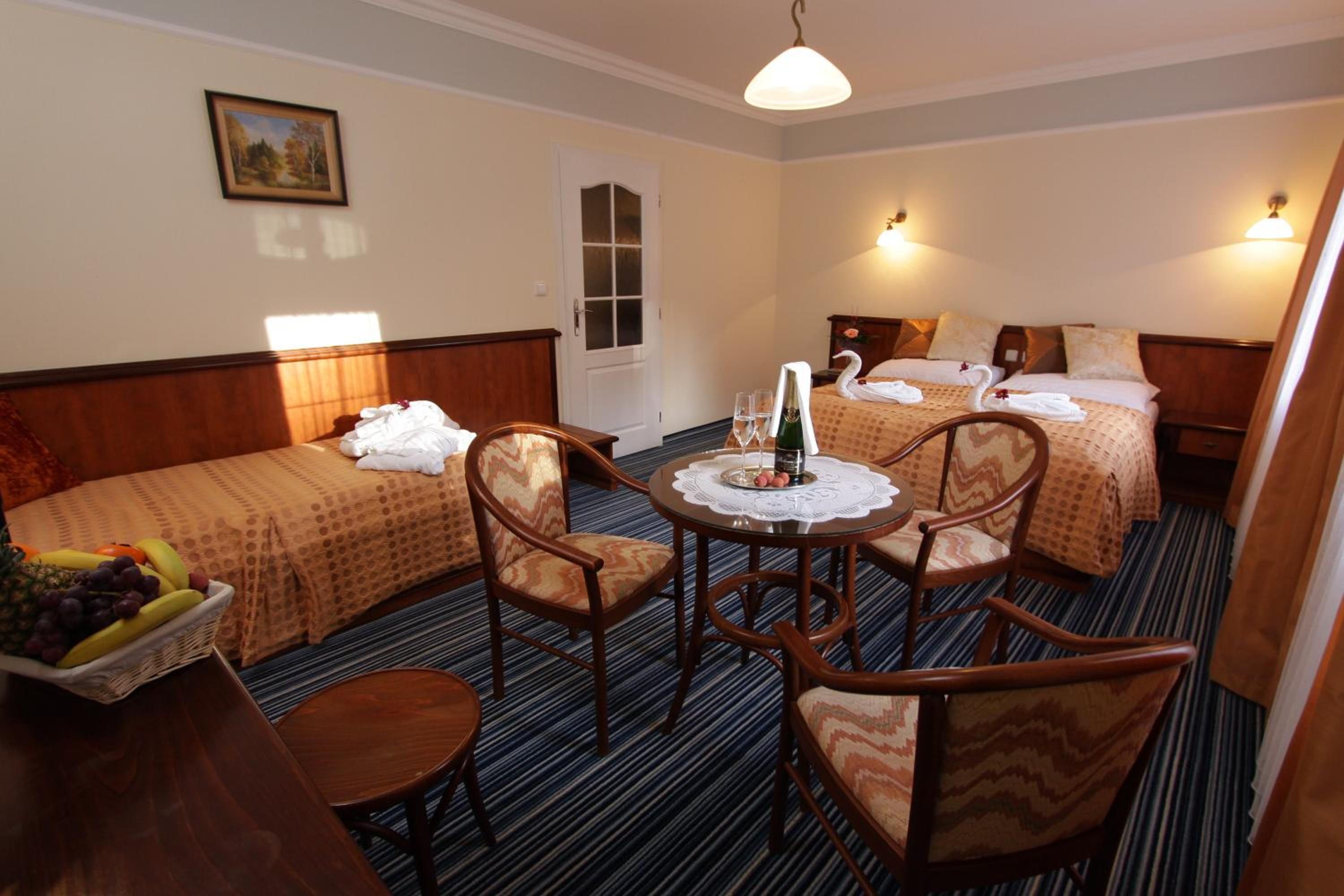 Habitación de hotel con dos camas individuales, una pequeña mesa de comedor con sillas, una cesta de frutas y un cuadro en la pared.