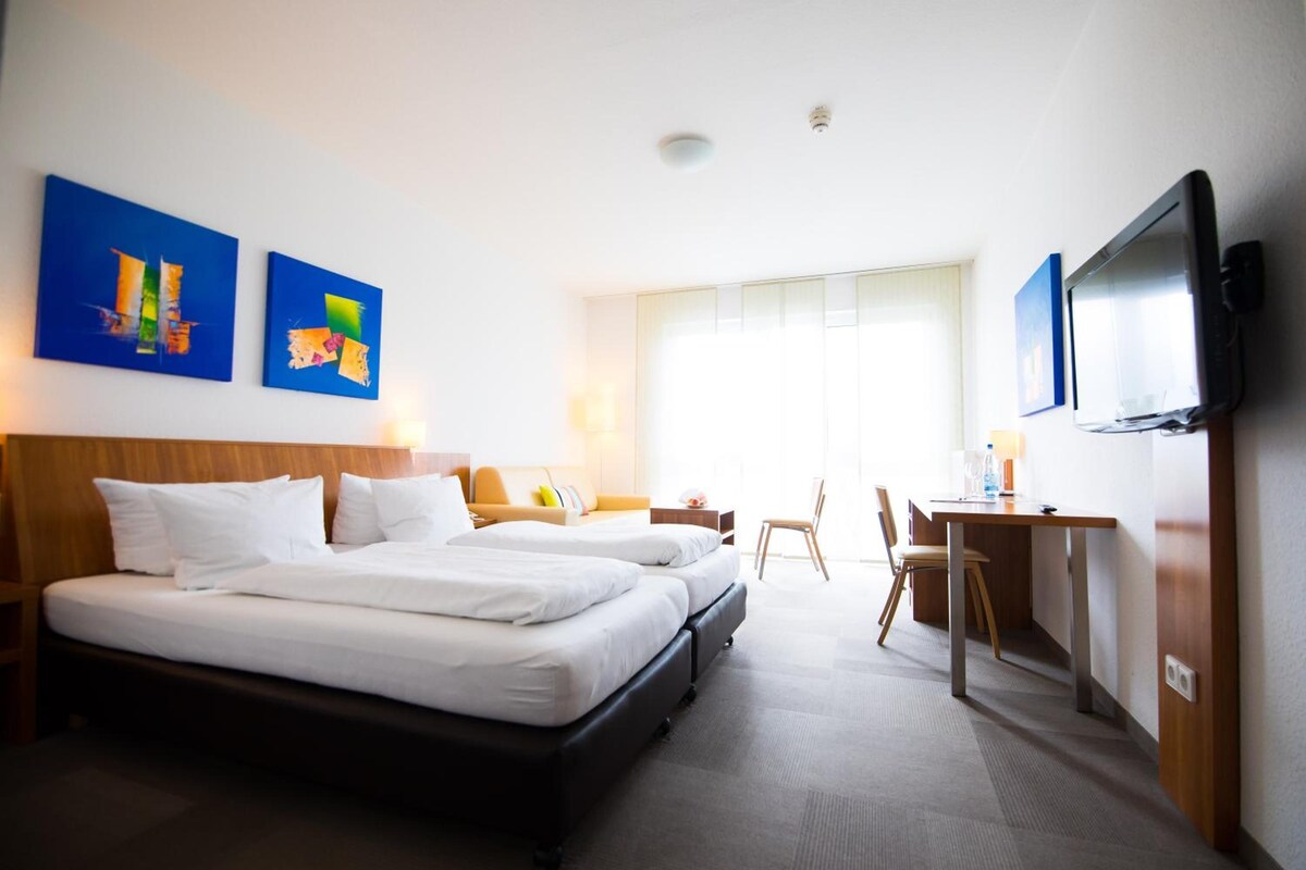 A hotel room with two beds, a desk, a chair, a television, and artwork on the walls.