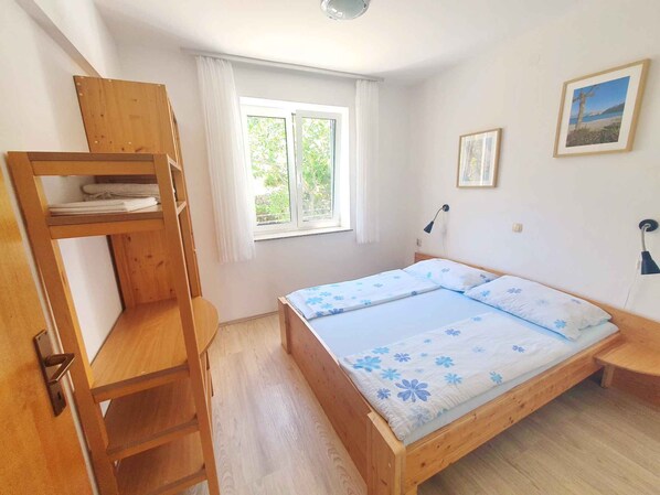 Une chambre avec un lit superposé en bois, un lit simple avec une literie à motifs floraux, une fenêtre avec des rideaux, une lampe fixée au mur et un tableau encadré.