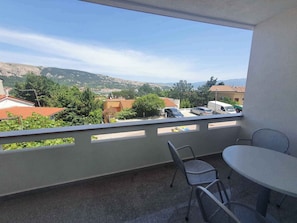 A balcony with a table and chairs, overlooking a residential area with houses and a hilly landscape.