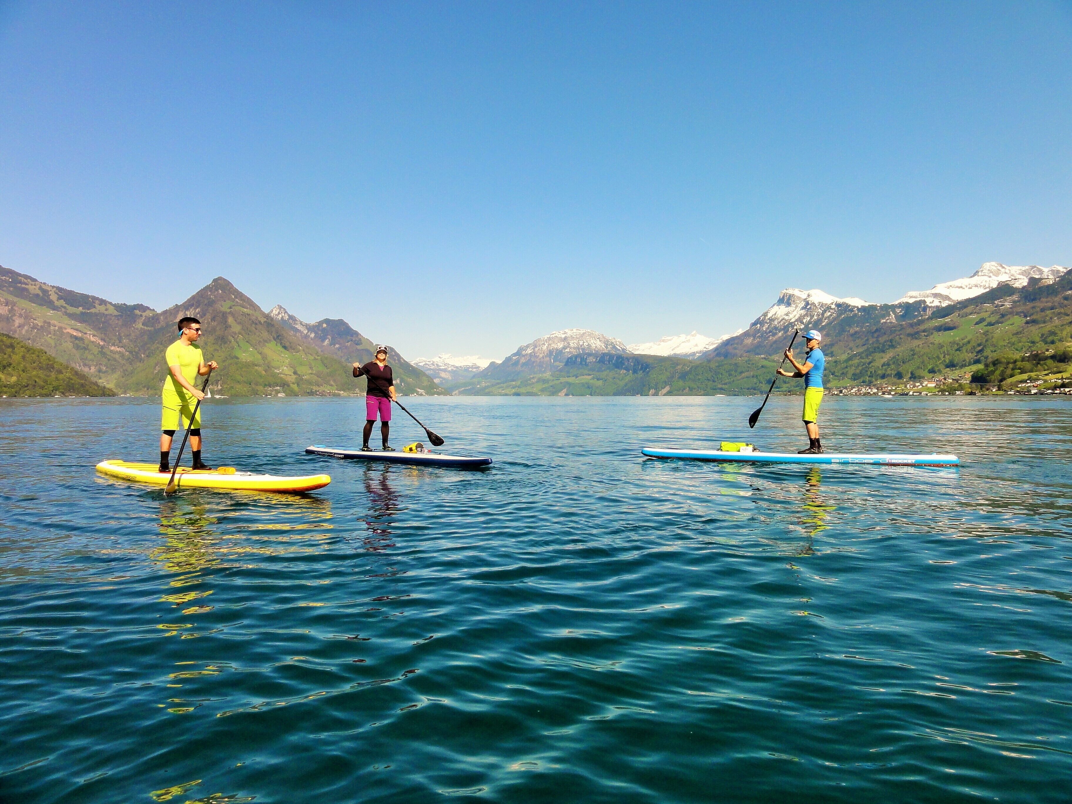 Trois personnes pratiquent le paddle sur un lac paisible, avec des montagnes en arrière-plan.