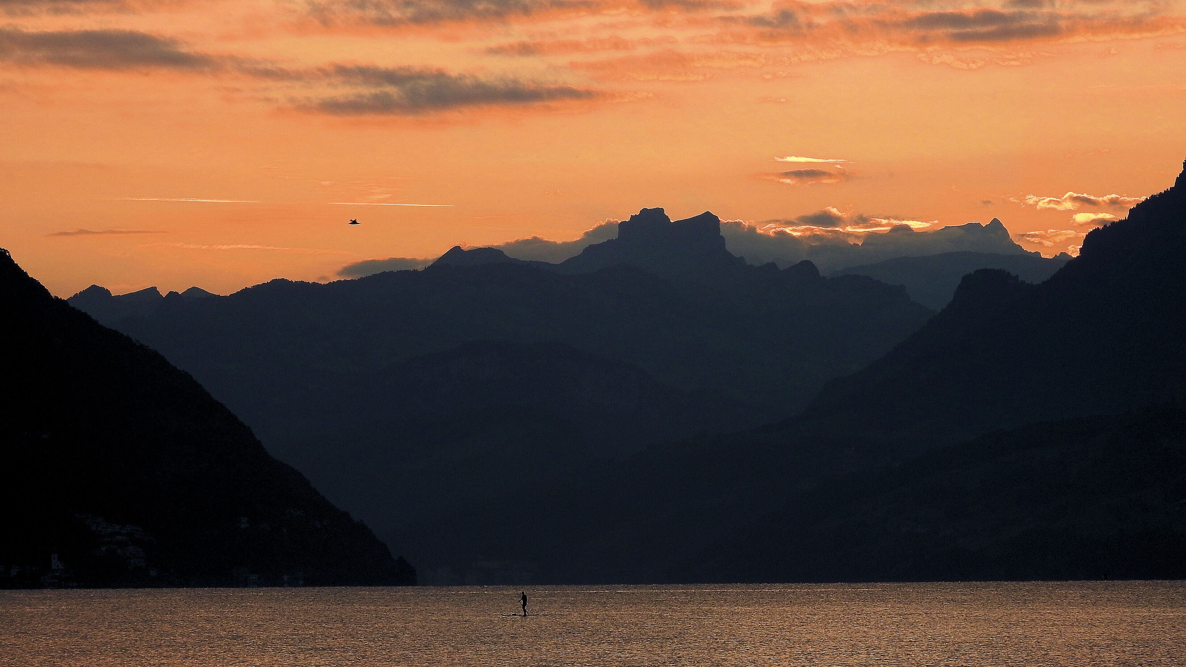 Un paysage montagneux où l’on voit une personne se tenant dans une étendue d’eau, probablement un lac ou la mer, au coucher du soleil.