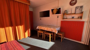 Een kamer met een bed, een tafel met stoelen, een televisie, een plank met decoratieve objecten en een radiator.