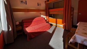 Een kamer met een enkel bed en een stapelbed op de achtergrond, een tafel met een bloemrijk tafelkleed, en een raam met gordijnen.