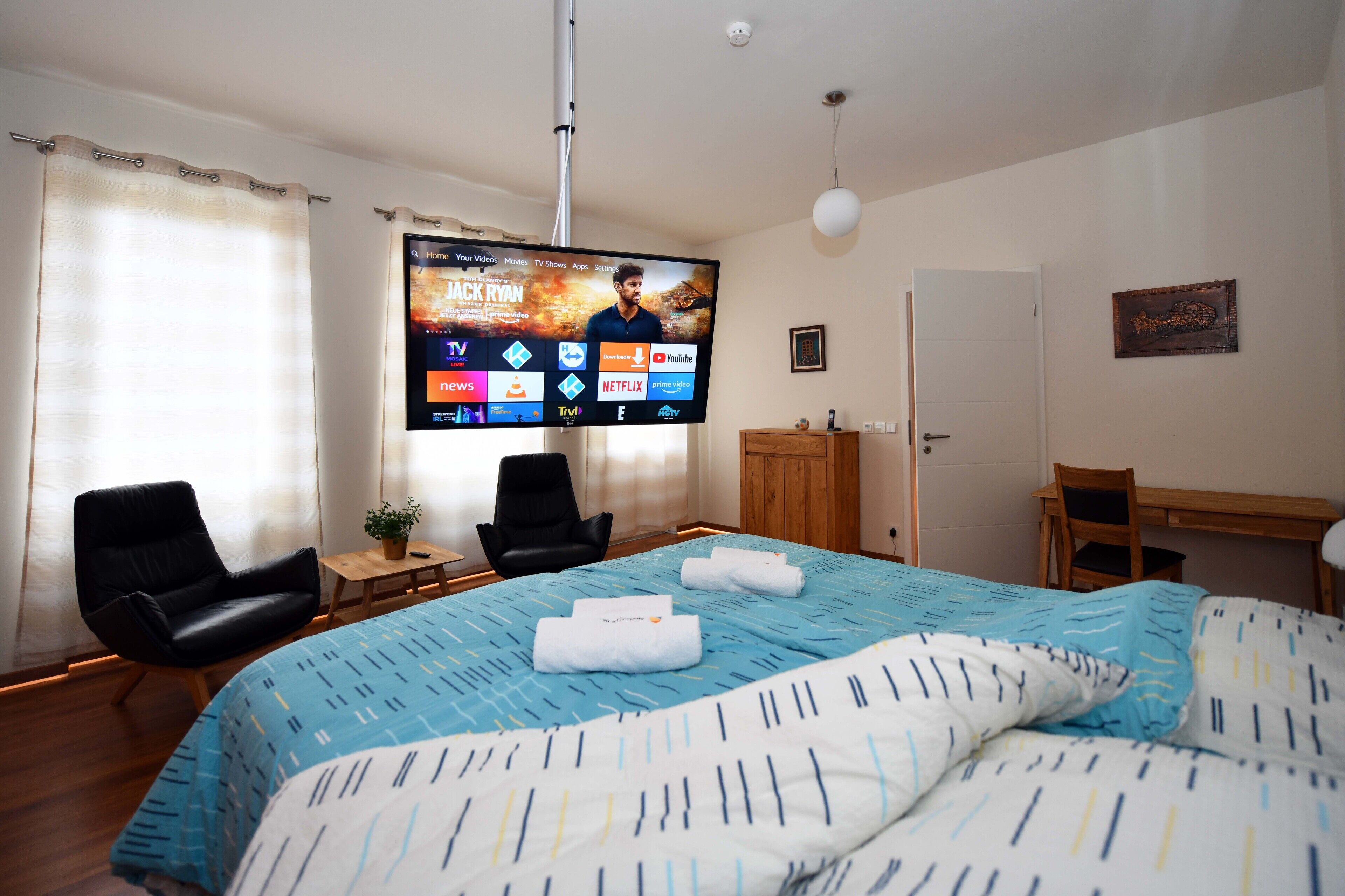 Ein Hotelzimmer mit einem großen Bett, zwei Sessel, einem Schreibtisch, einem flachbildfernseher an der Wand und einer Tür.