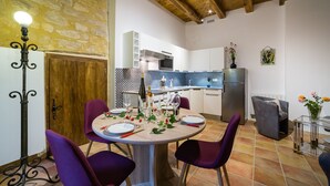 Un comedor con una mesa redonda dispuesta para cuatro personas, rodeada de sillas moradas. Se ve una cocina con electrodomésticos modernos y un salpicadero a cuadros al fondo.