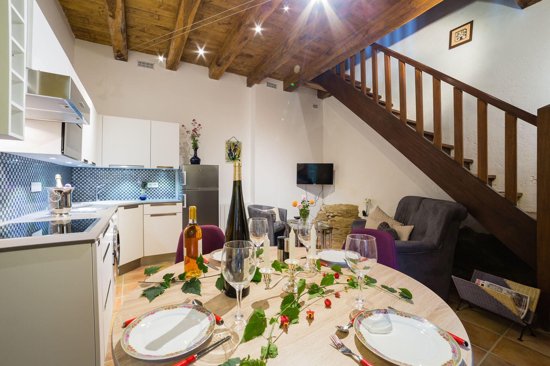 Een eethoek met een tafel voor vier personen, een keuken met een zwart-wit geruit spatwand, en een trap naar een hoger niveau.
