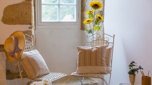 Una habitación acogedora con ventana, una silla de metal, un cojín, un sombrero y un jarrón con girasoles.