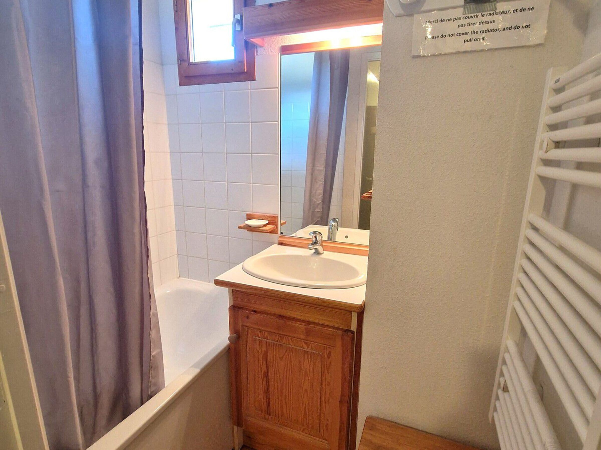 A small bathroom with a bathtub, a sink, and a mirror. There is a window with curtains and a sign on the wall.