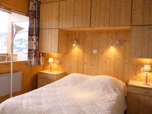 A room with wooden panel walls, a bed with a white quilt, two bedside tables with lamps, and a window with a curtain. - Cozy 2-room apartment in Méribel, 1 bedroom, for 3 people (Méribel)