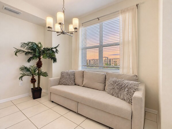 A living room with a beige sofa, a potted plant, and a window with blinds.
